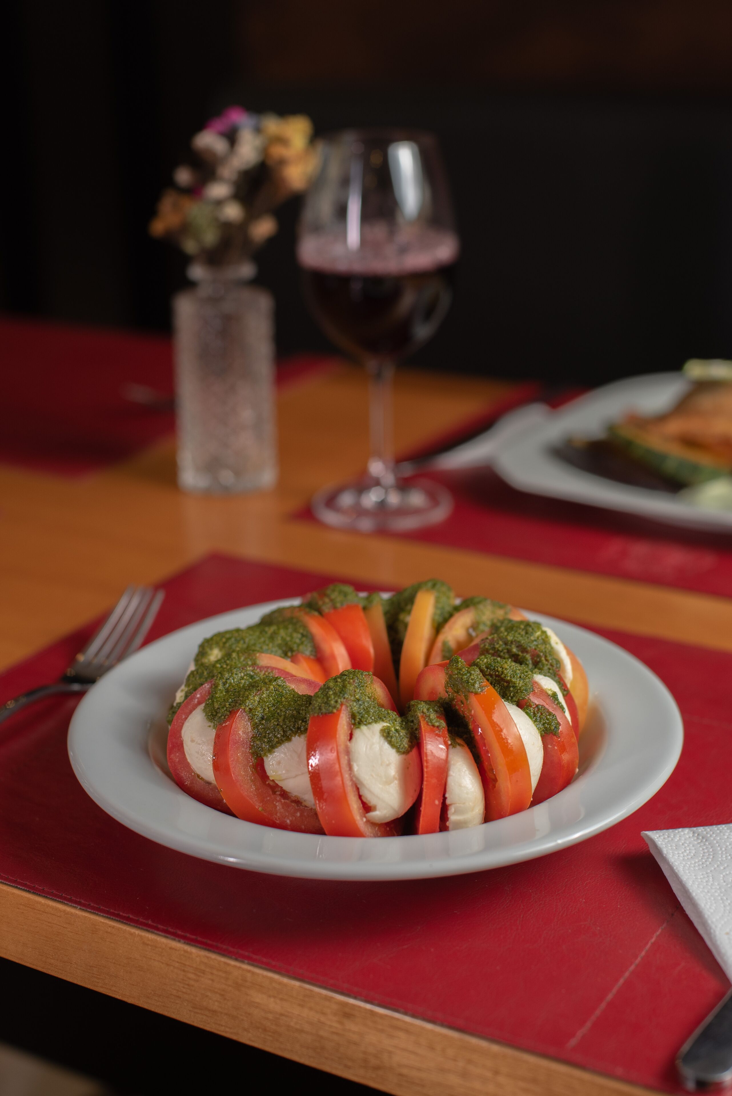A plate of Caprese salad with tomatoes, mozzarella, and basil, a glass of red wine, and a vase of flowers on a table.