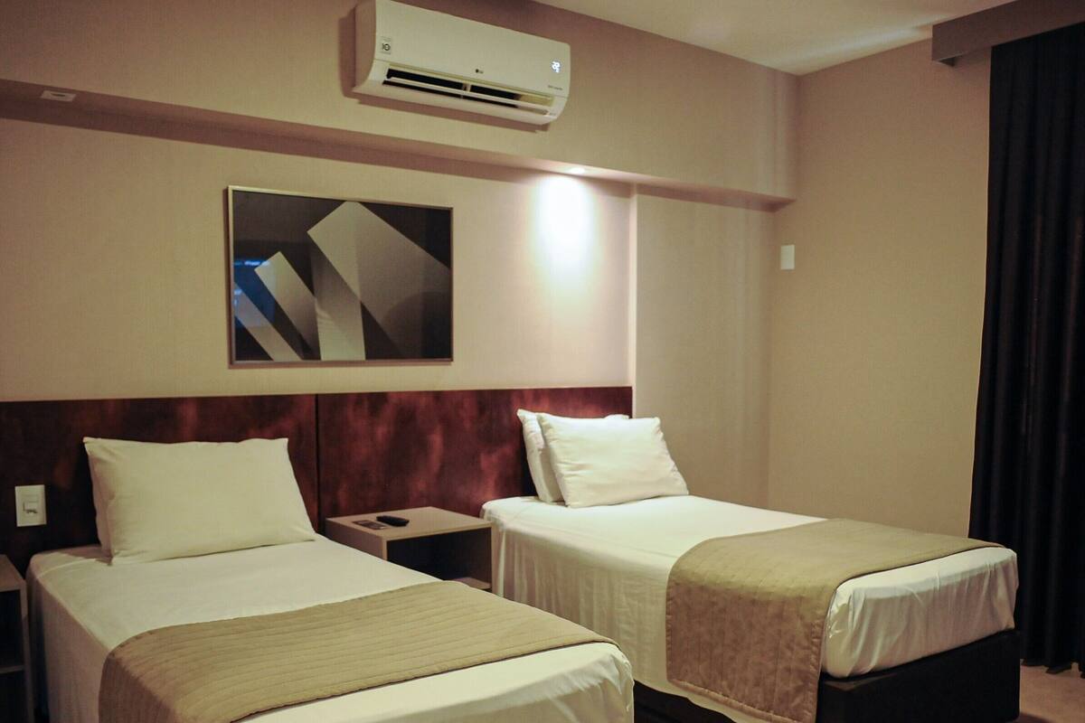 A hotel room with two beds, a nightstand, a wall-mounted air conditioner, and a painting.