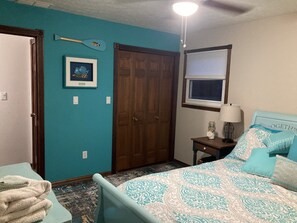 A bedroom with teal walls, a bed with patterned bedding, a wooden door, a framed picture, a window with blinds, and a ceiling fan.
