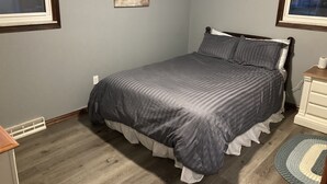 Een slaapkamer met een bed, een nachtkastje, een raam en een schilderij aan de muur.