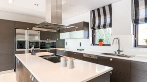 A modern kitchen with a central island, stainless steel appliances, and a window with striped curtains. - East Coast Mansion (Ανάβυσσος)