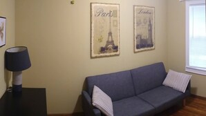 A living room with a grey sofa, two framed pictures of Paris and London, a lamp, and a window.