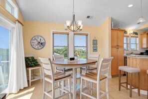 A dining area with a round table, white chairs, and a chandelier. There is a window with blinds, a potted plant, and a wall clock. - The Dancing Sea Horse: Private Pool, Elevator, Spa (Kitty Hawk)