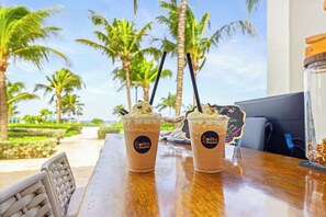 Dos cafés helados con crema batida y virutas de chocolate sobre una mesa de madera, con vista a palmeras y una playa.