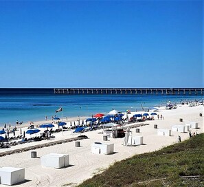 A beach with rows of sunbeds and umbrellas, a pier in the background, and a clear blue sky.