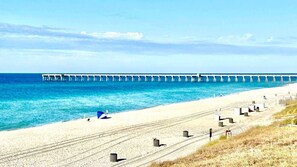 Une plage avec une jetée qui s’avance dans l’océan, un sentier de sable et des poubelles disposées le long du chemin.