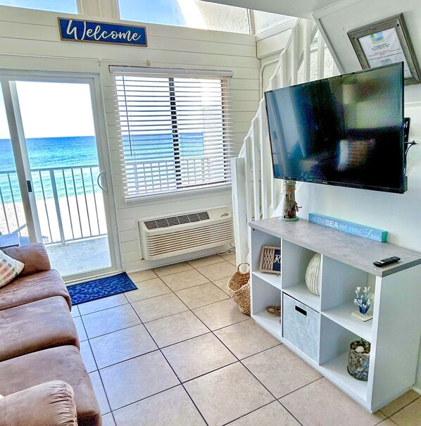 A living room with a TV, a 'Welcome' sign, a view of the ocean, a sofa, and a small shelf with decorative items.