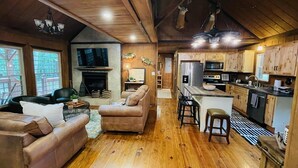 A cozy cabin interior with a living room, fireplace, kitchen, and wooden ceiling.