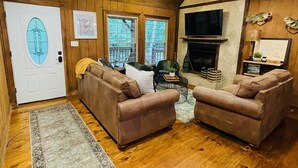 A cozy living room with wooden floors and walls, two sofas, a chandelier, a fireplace, and a TV mounted on the wall.