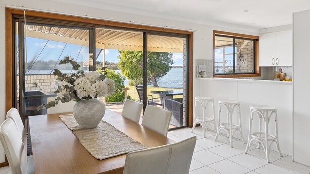 A dining area with a table, chairs, a vase of flowers, and a view of a lake through large glass doors. - Peninsula Court Unit 10 - Waterfront Unit with Pool & River Views - Holiday Yamba (YAMBA)