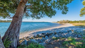 A beach with a rocky shoreline, a clear blue sky, and a tree in the foreground. - Peninsula Court Unit 10 - Waterfront Unit with Pool & River Views - Holiday Yamba (YAMBA)