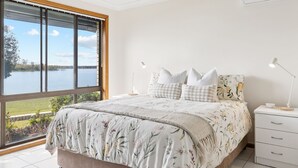 A bedroom with a large window, a bed with floral bedding, two bedside lamps, and a white dresser. - Peninsula Court Unit 10 - Waterfront Unit with Pool & River Views - Holiday Yamba (YAMBA)