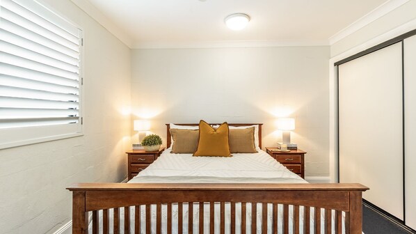 A bedroom with a wooden bed frame, two bedside tables with lamps, a nightstand with a plant, and a window with blinds. - Arcoola 2 - Views Over Main Beach - Holiday Yamba (Yamba)