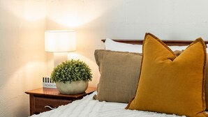 Un lit bien fait, avec une couette blanche, un oreiller de couleur moutarde et un oreiller marron posés sur une table de chevet en bois. On peut également apercevoir une petite plante en pot et un panneau décoratif sur le thème nordique.