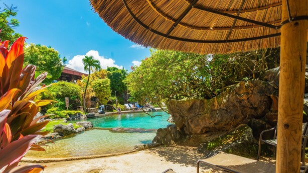 Zona de piscina con sillones de descanso, una sombrilla de paja y un telón de fondo rocoso.