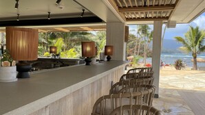 Zona de bar con sillas de madera y una barra, con vistas a una playa con palmeras y el océano.
