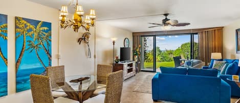 Amplia sala de estar con sofá azul, sillas de mimbre, mesa de comedor de vidrio y ventilador de techo.