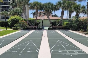 A shuffleboard court with a green surface, surrounded by palm trees and a building in the background.