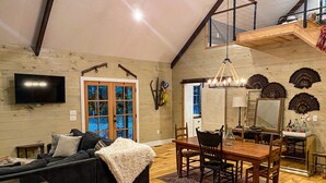 A cozy living room with a black sofa, wooden dining table, and a TV mounted on the wall. - Quaint Little Cabin with a view. (Abbeville)