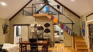A spacious loft with a high ceiling, exposed wooden beams, a dining area with a large wooden table and chairs, a staircase with colorful ribbons, and a cozy bedroom with a bed and nightstands. - Quaint Little Cabin with a view. (Abbeville)