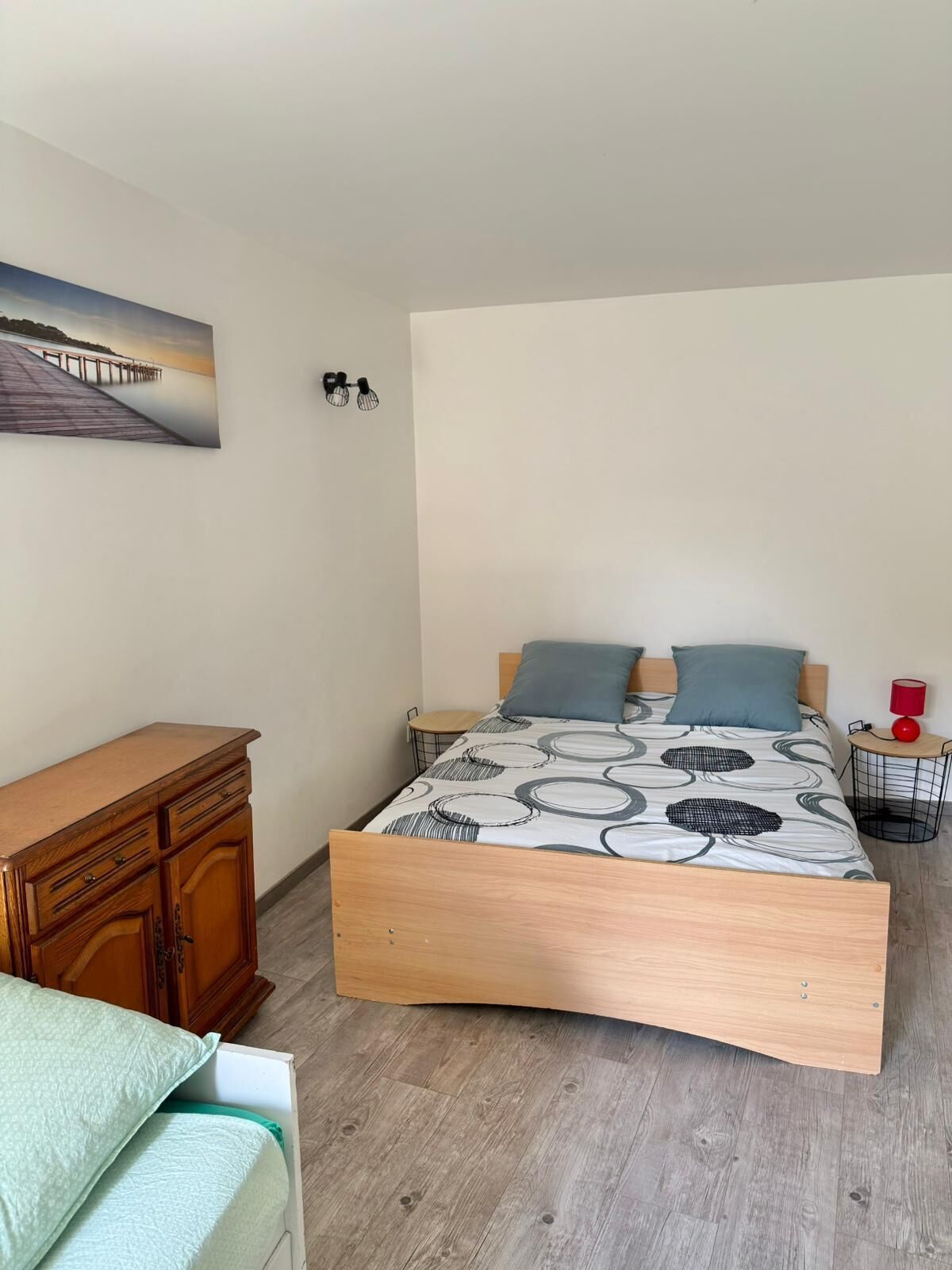 Een slaapkamer met een houten bed, een houten dressoir, een tafeltje en een wandlamp.