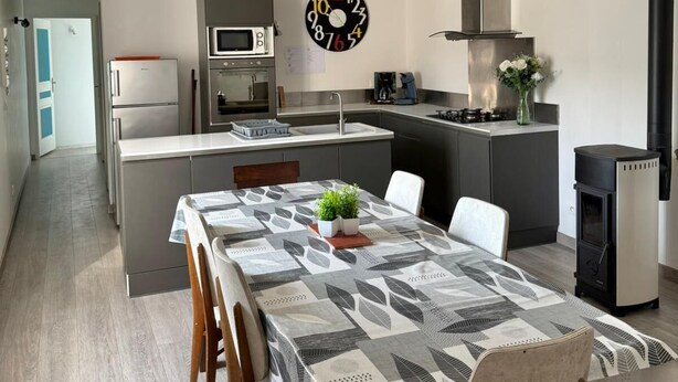 Een moderne keuken met een centraal eiland, een eetgedeelte met een tafelkleed met patroon en een grote wandklok.