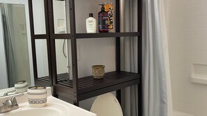A bathroom with a white toilet, a wooden cabinet, a black shelf unit with toiletries, a white sink, a mirror, a fan, and a shower curtain. - Private Room on Serene Property in Hilo (Hilo)