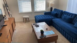 Ein Wohnzimmer mit einem blauen Sofa, einem Holztisch mit Papieren, einem kleinen Hocker und einem Fernseher auf einer Holzkommode.