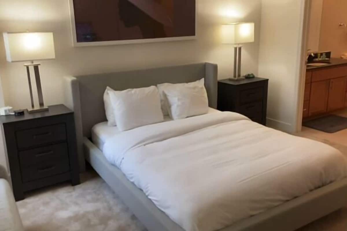 A neatly made bed with white linens, two bedside tables with lamps, a grey headboard, and a small painting above the bed.