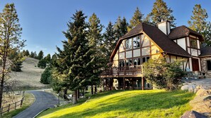 Une maison à deux étages avec un balcon, entourée d’arbres et bordée d’une pente herbeuse.