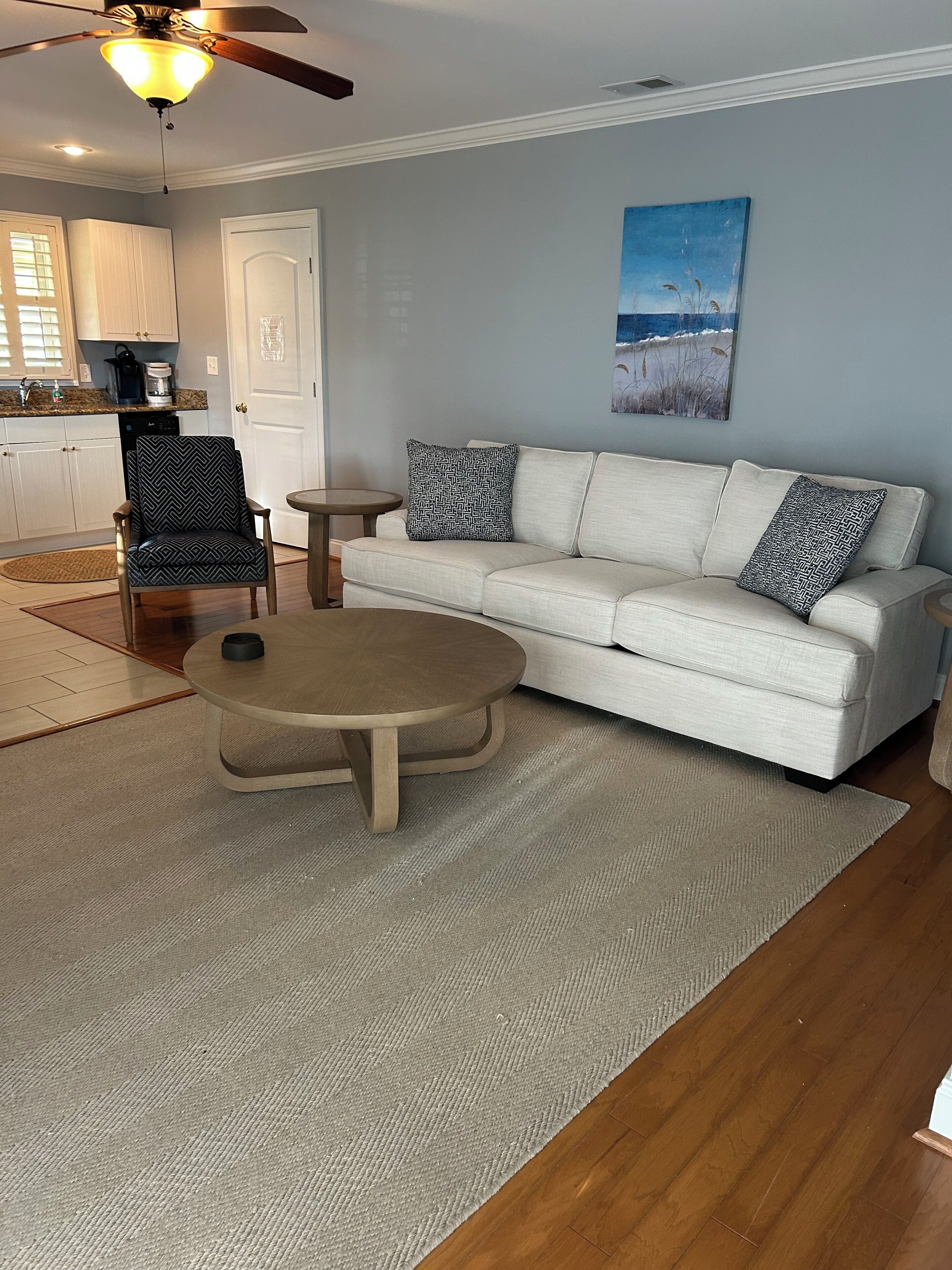 Un salon avec un canapé beige, des coussins à motifs, une table basse ronde, un fauteuil à motifs et un tableau accroché au mur.