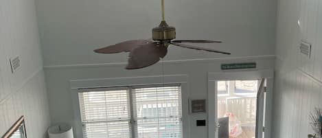 A spacious living room with a high ceiling, a ceiling fan, a light blue sofa, a matching armchair, and a large window with blinds.