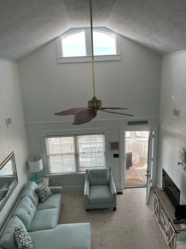 A spacious living room with a high ceiling, a ceiling fan, a light blue sofa, a matching armchair, and a large window with blinds.
