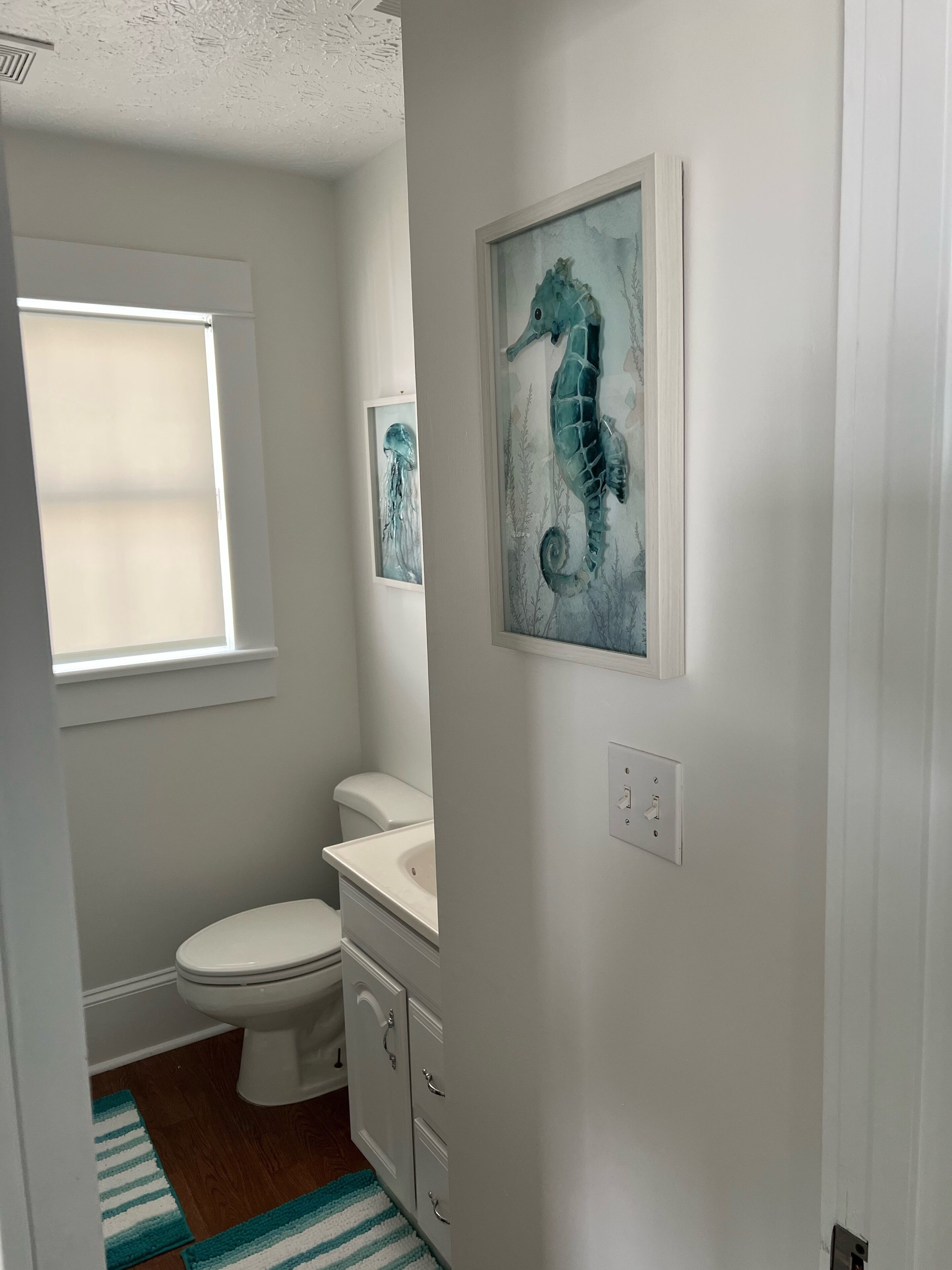 Ein Badezimmer mit Toilette, Waschbecken und einem Fenster. Zwei gerahmte Kunstwerke befinden sich an der Wand.