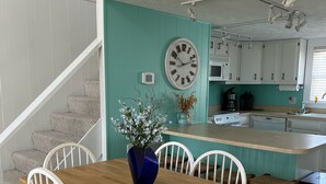 Un comedor con mesa de madera y sillas blancas, un jarrón azul con flores, un reloj en la pared y una cocina al fondo.