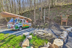 A colorful bus parked at a campsite with a large canoe on top, surrounded by rocks and trees.