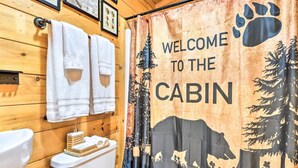 Baño de cabaña con paredes de madera, inodoro, lavamanos y cortina de ducha con diseño de oso y bosque.