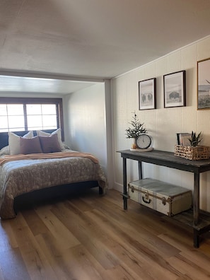 A bedroom with a bed, a wooden floor, a small table with a clock, and framed pictures on the wall.