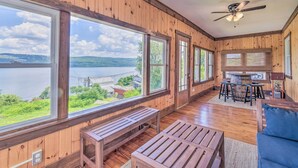 A wooden cabin with large windows overlooking a lake, a ceiling fan, and a dining area with a table and chairs. - Watkins Glen Lakeview Cottage w/ Waterfall! (Watkins Glen)