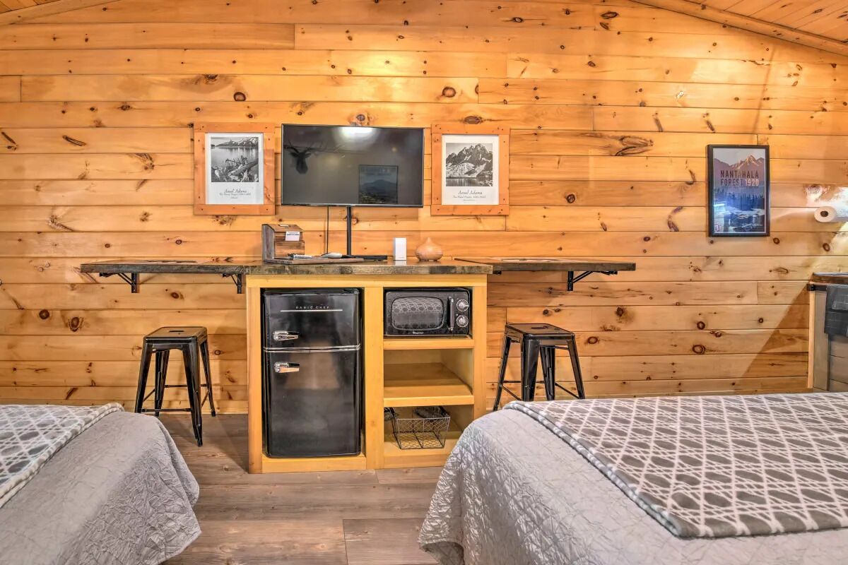 Ein gemütliches Blockhauszimmer mit Holzwänden, einer zentralen Küchenzeile, einem Flachbildfernseher und zwei Betten mit gemusterten Betttextilien.