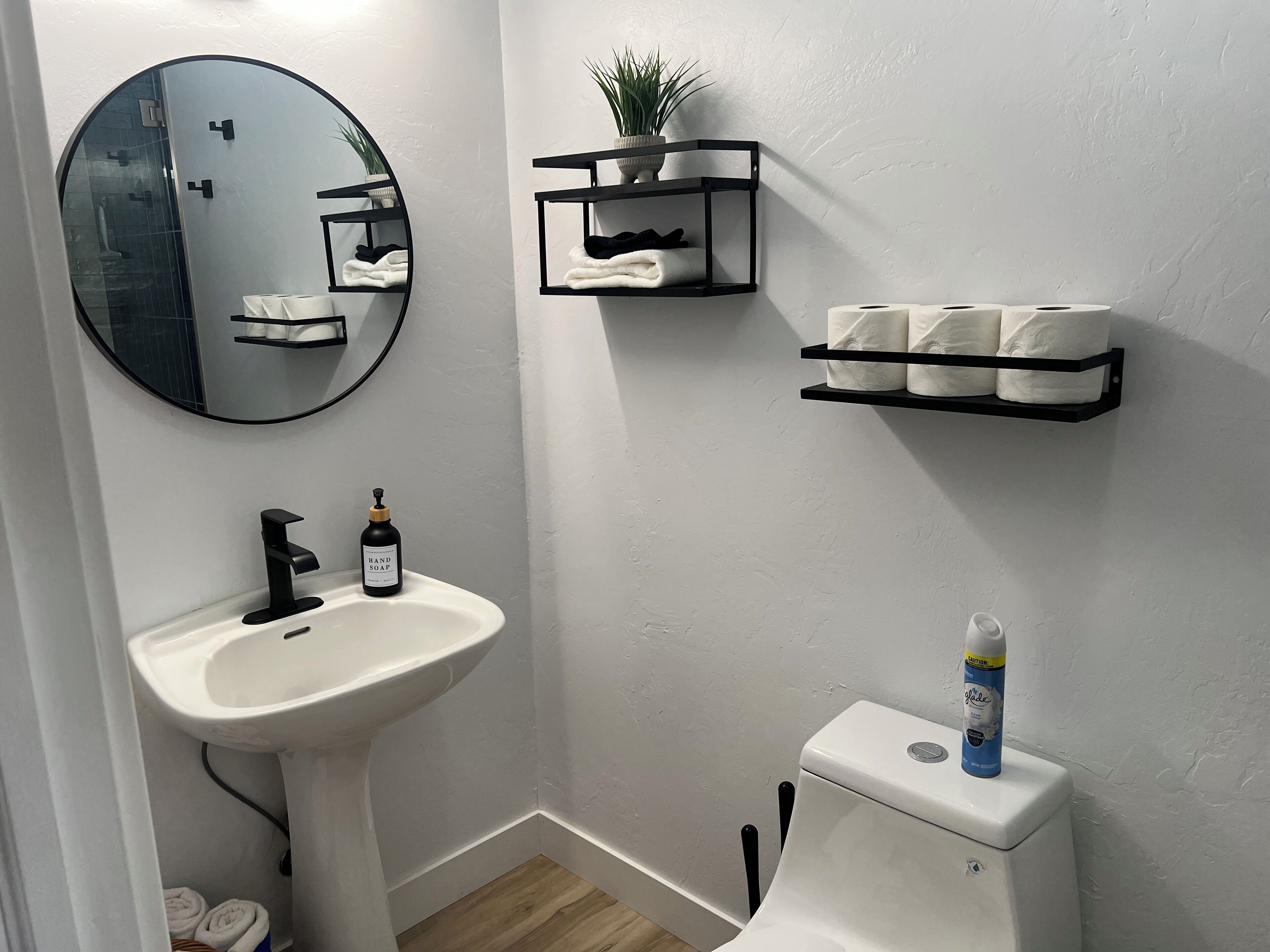 Une salle de bain avec un lavabo blanc, un miroir rond, des toilettes et des étagères contenant des serviettes et des articles de toilette.