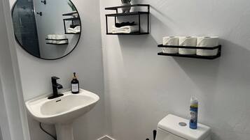 Une salle de bain avec un lavabo blanc, un miroir rond, des toilettes et des étagères contenant des serviettes et des articles de toilette.