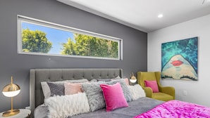 Ein modernes Schlafzimmer mit einem großen Bett, Blick auf Bäume durch ein Fenster, einem Bild an der Wand und einem gelben Sessel.
