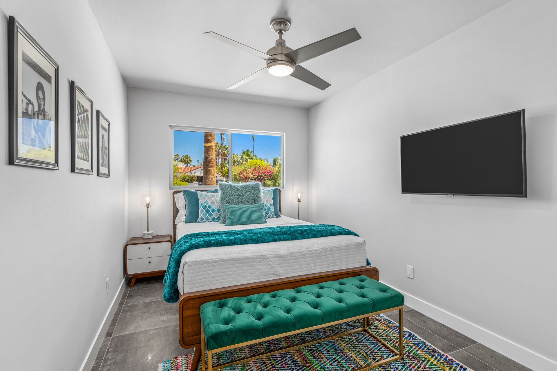 Een slaapkamer met een bed, een plafondventilator, een raam met uitzicht op palmbomen, een aan de muur bevestigde televisie en een kleurrijk vloerkleed.