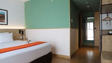 A hotel room with a bed, a wooden headboard, a green accent wall, a wooden floor, a hallway with a door, and a hanging coat rack.