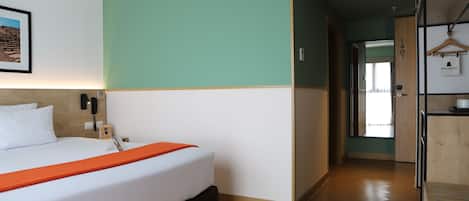 Una habitación de hotel con una cama, un cabecero de madera, una pared de acento verde, piso de madera, un pasillo con una puerta y un perchero.