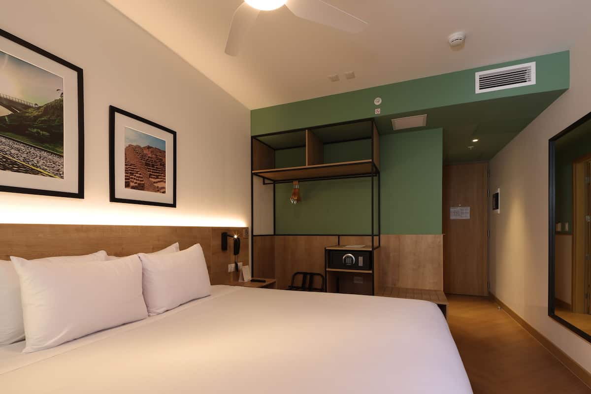 A hotel room with a bed, a desk, and a bunk bed. There are framed pictures on the wall, a ceiling fan, and an air conditioning unit.