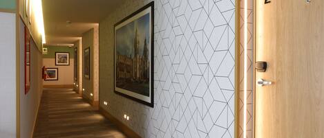 Un couloir d’hôtel avec un mur orné d’un motif géométrique, des tableaux encadrés et une porte en bois portant le numéro 302.