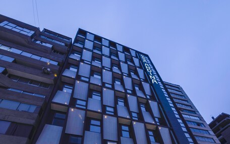 A modern hotel with a glass facade, multiple floors, and a well-lit entrance with steps leading up to the doors.
