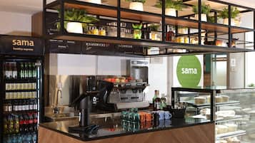Un café con una máquina de café, estanterías con bebidas y una vitrina con pasteles.